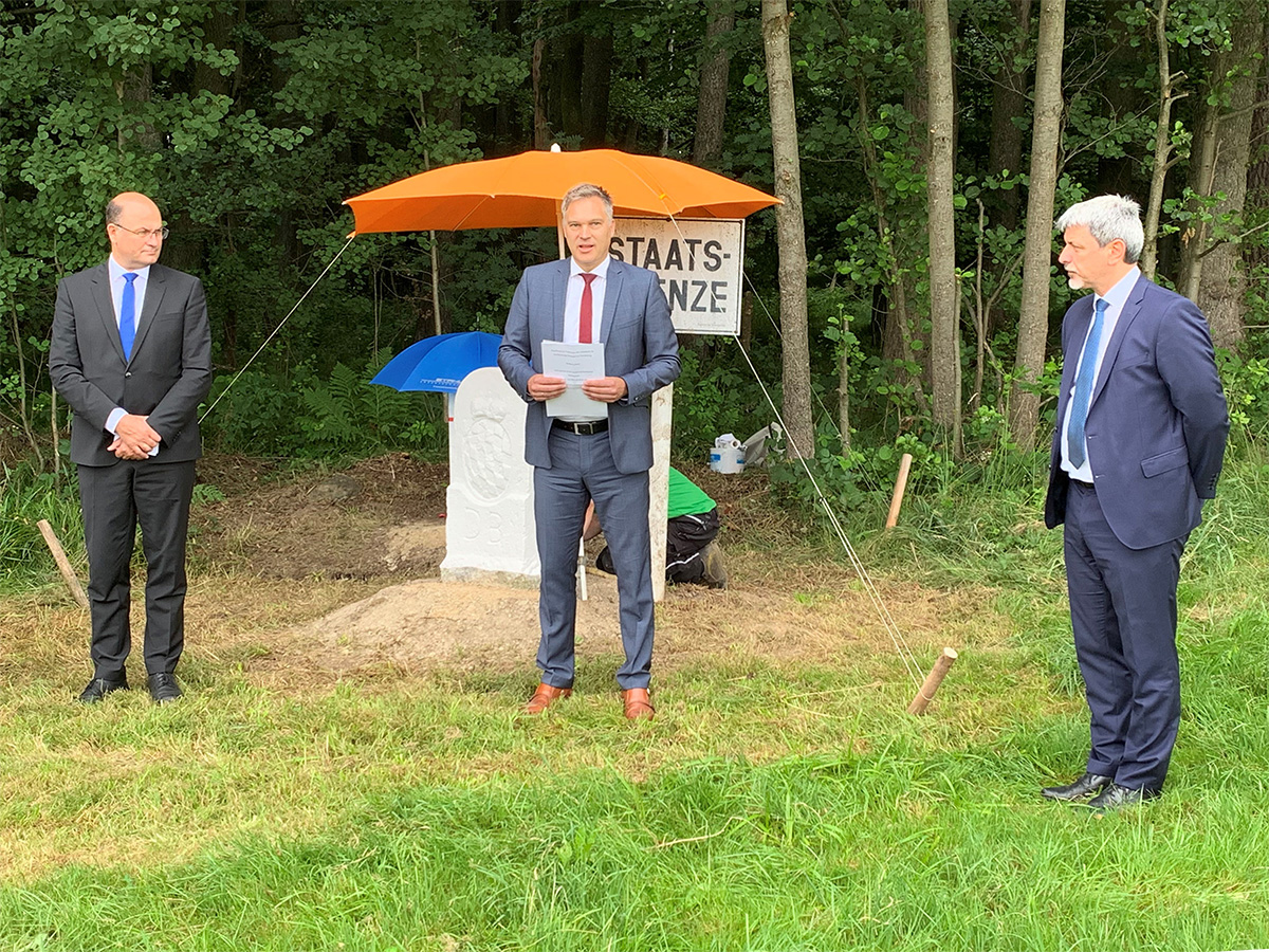 Wolfgang Bauer, flankiert von zwei weiteren Männern, hält eine Rede vor dem historischen Grenzstein. Die Namen der abgebildeten Personen stehen in der Bildunterschrift.
