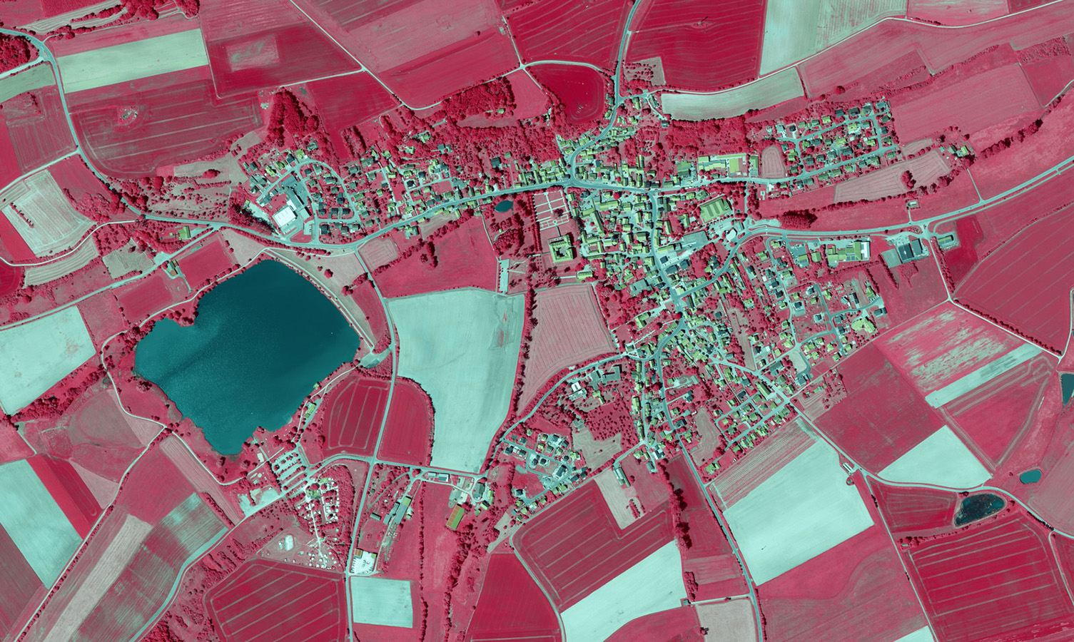 Das CIR-Luftbild zeigt Vegetation in roter Farbe, Gewässer in Blau und Gebäude in Gelb und Grau. Zu sehen ist ein See, Wiesen und Felder sowie eine Siedlung.