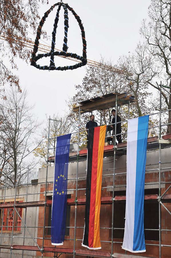 Vor dem Rohbau hängen drei Fahnen, die EU-Fahne, die Fahne der BRD und die Fahne Bayerns. Über den Fahnen schwebt eine Richtfestkrone.